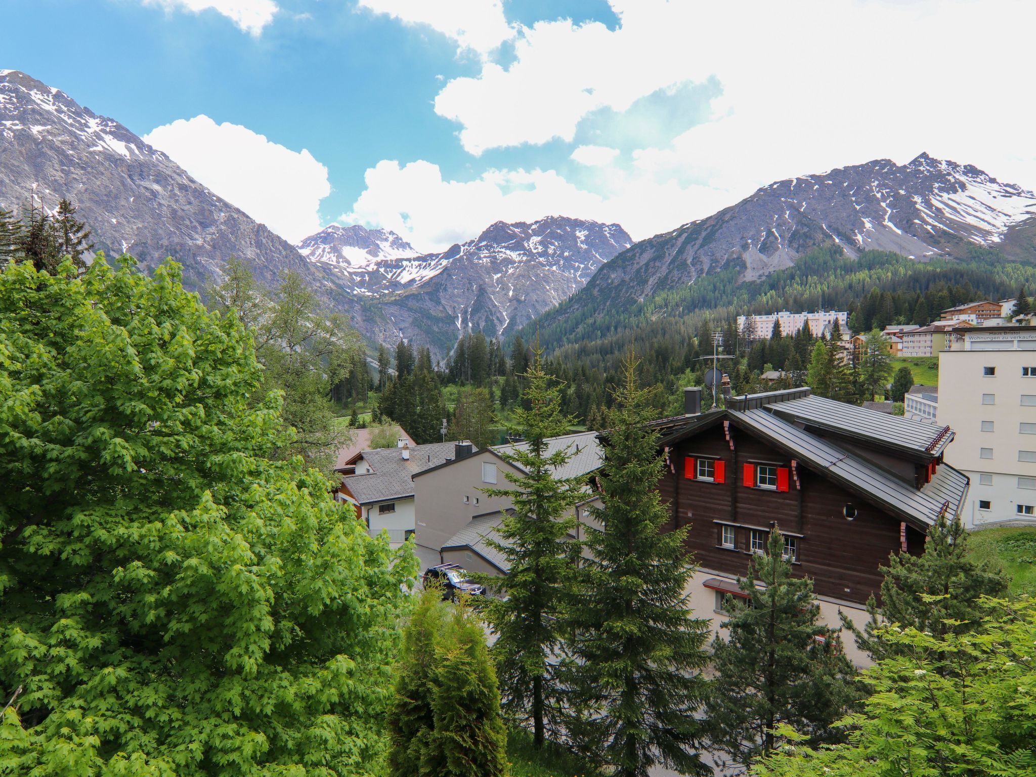 A mountain town with traditional wooden houses, a modern building, and snow-capped peaks in the background.