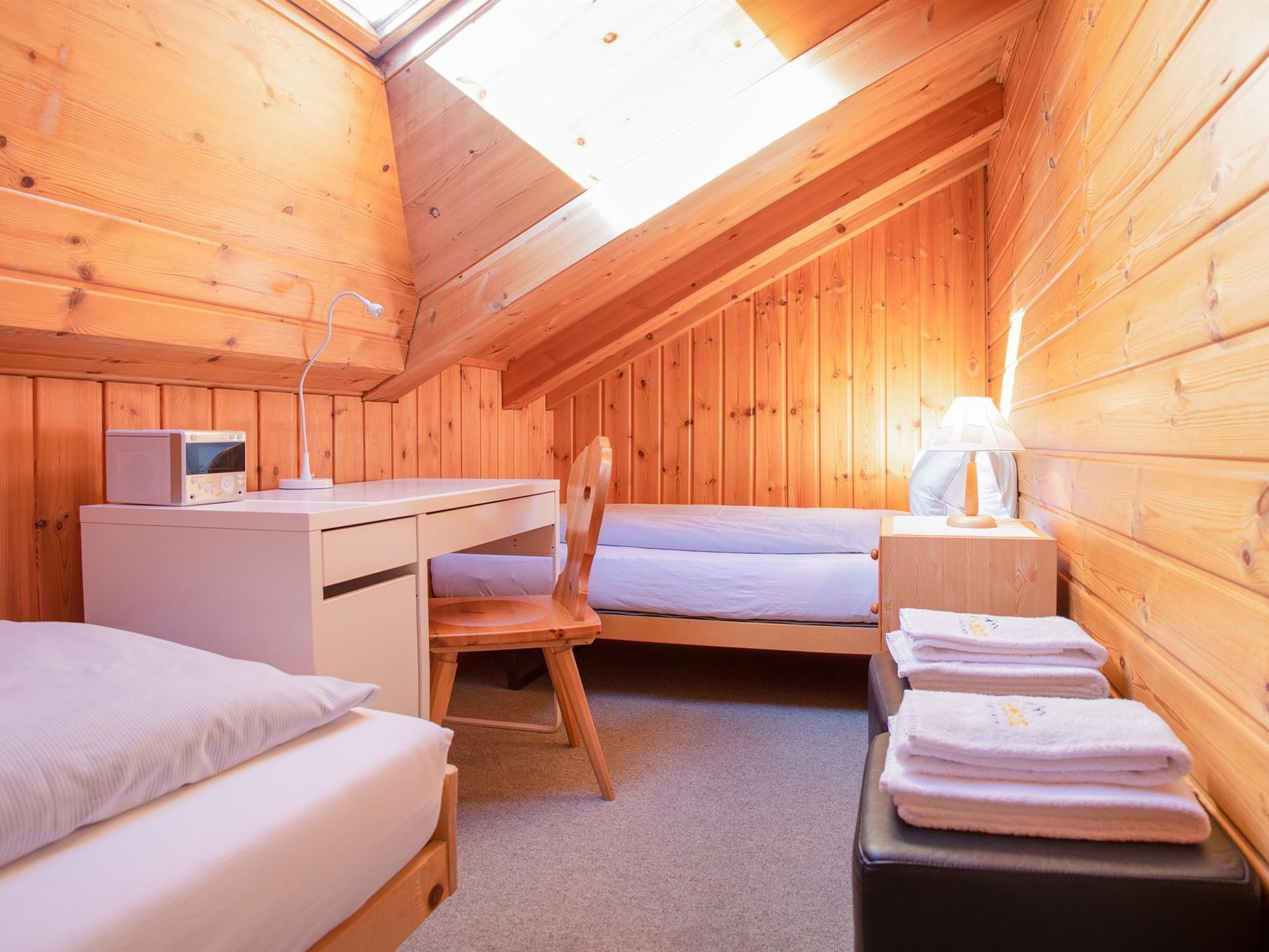 Um quarto pequeno e acolhedor com paredes de madeira e teto inclinado, com uma cama, uma escrivaninha com computador e uma cadeira.