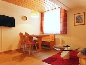 A cozy room with a wooden table, chairs, a glass-top coffee table, a television, a clock, and a window with curtains. - Furkablick Yvonne by Arosa Holiday Appartement/Fewo, Bad, WC Furkablick Yvonnne by Interhome (Arosa)