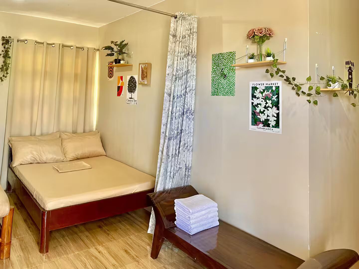 Una camera da letto con un letto, un comodino in legno, una finestra con tende e decorazioni alle pareti tra cui un quadro incorniciato e una piccola mensola con piante.