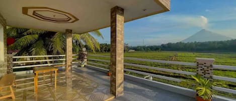 A covered terrace with a view of a mountain and rice fields.