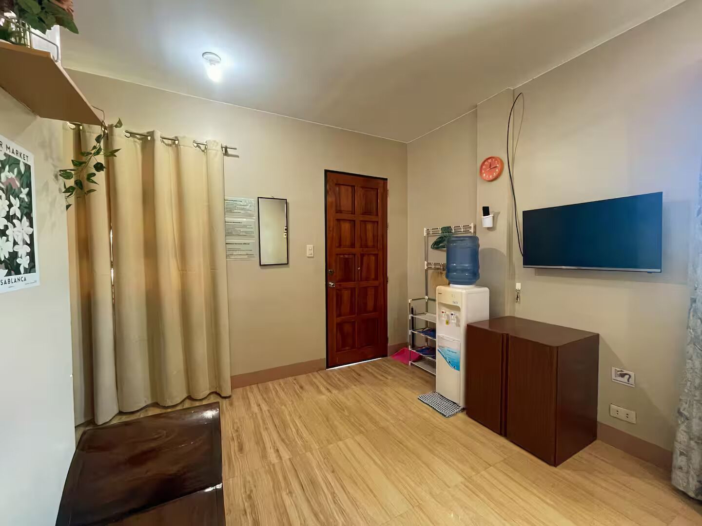 A living room with a wooden floor, a television mounted on the wall, a water dispenser, and a wooden cabinet.