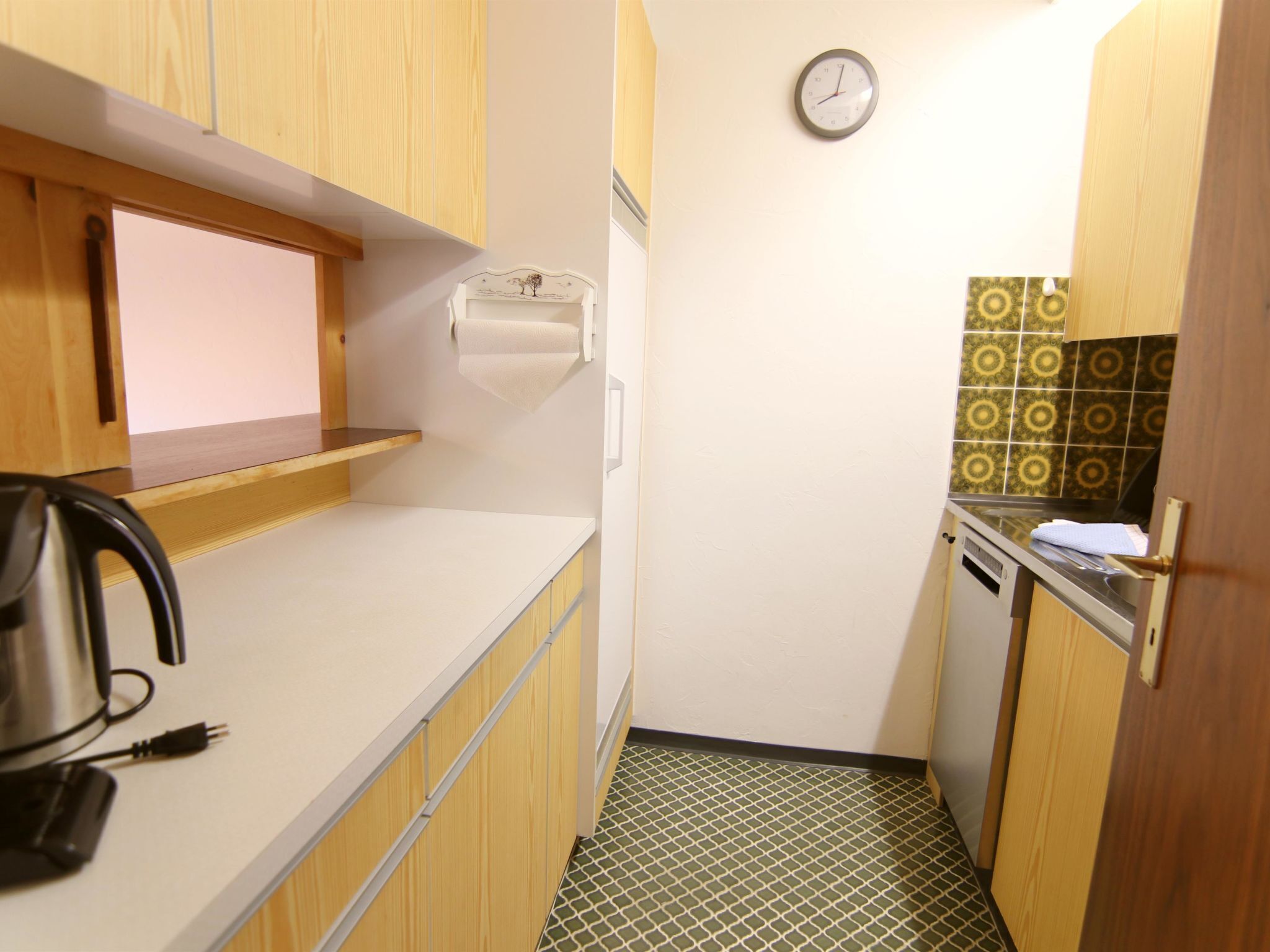 A compact kitchen with wooden cabinets, a sink, a clock, and a patterned backsplash.