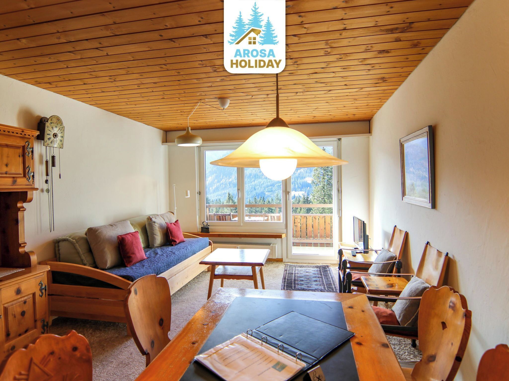 A cozy room with a wooden ceiling, a sofa, a dining table, and a large window offering a mountain view.