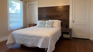 Ein Schlafzimmer mit Holzkopfstütze, einem Bett mit weißer Bettwäsche, einem Nachttisch, einem Fenster mit Vorhängen und einer Tür.