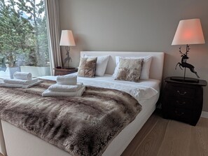 A hotel room with a large bed, two bedside tables with lamps, a fur throw, and a view of trees through the window.
