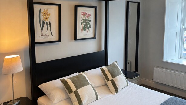 Habitación de hotel con cama, lámpara de noche, espejo en la pared e impresiones botánicas enmarcadas.