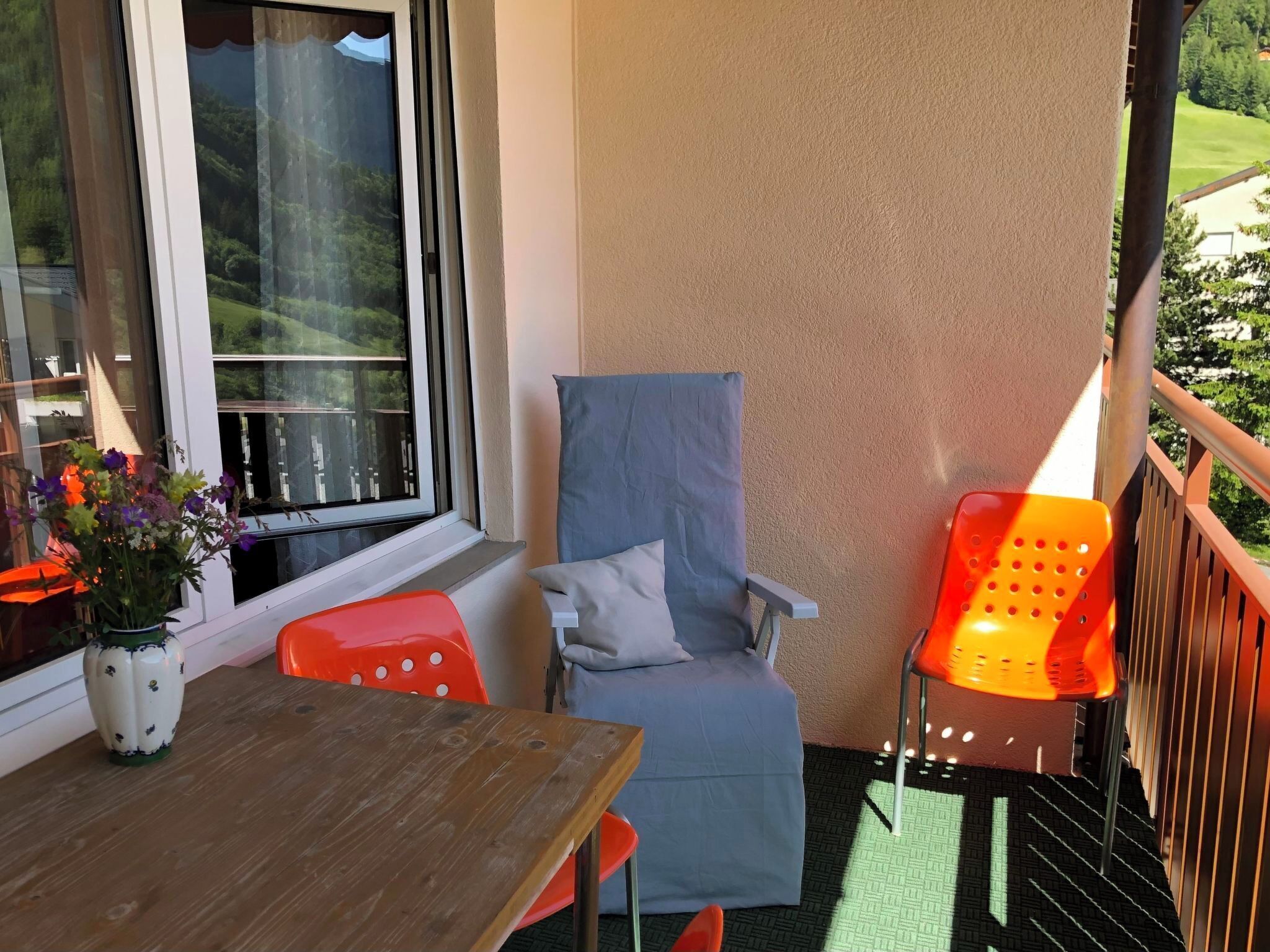 Ein Balkon mit Holztisch, zwei Stühlen, einem Topfpflanzen und Blick auf eine grüne Landschaft.