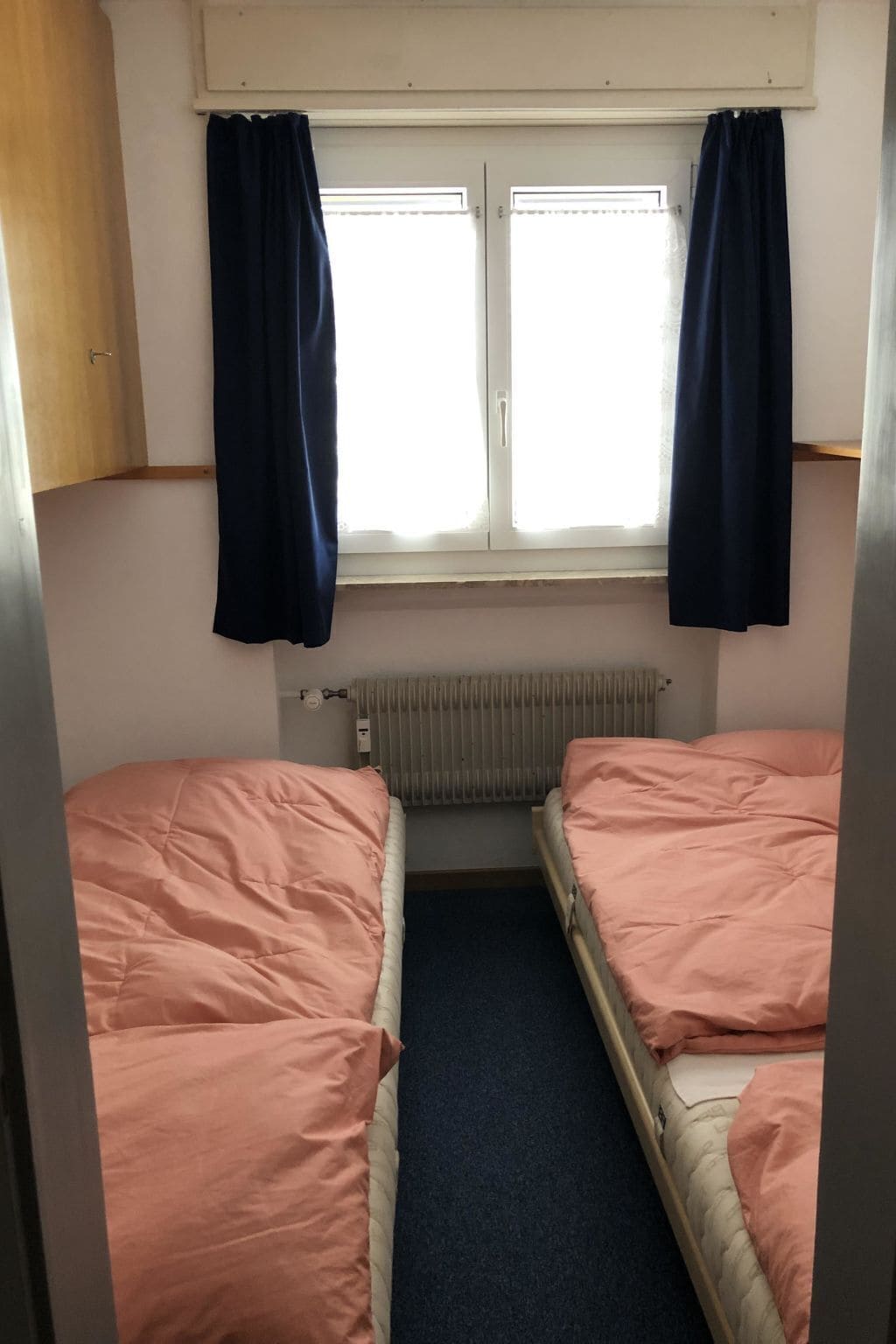 Une petite pièce avec deux lits, une fenêtre avec des rideaux, un radiateur et une armoire en bois.