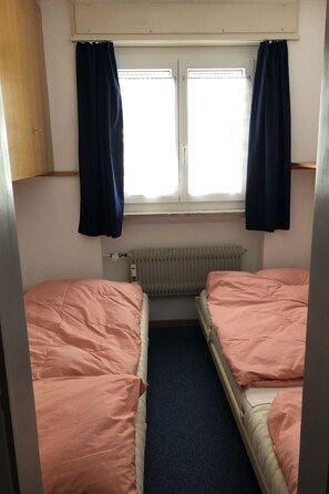 Ein kleiner Raum mit zwei Betten, einem Fenster mit Vorhängen, einem Heizkörper und einem Holzschrank.