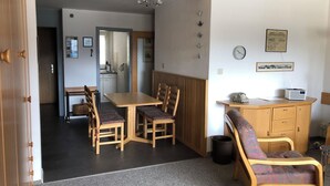 Un espacio habitable compacto con comedor, kitchenette y una pequeña zona de estar con sofá y sillones.