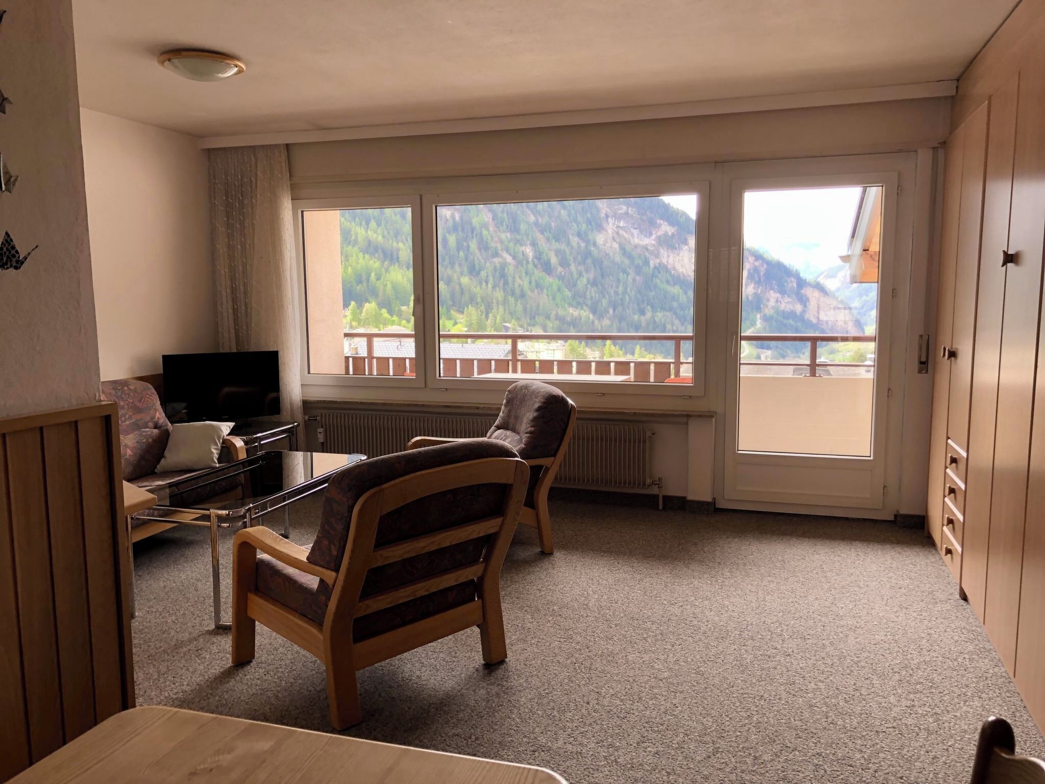 A room with a large window, a television, a wooden chair, and a table.