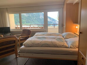 Habitación de hotel con cama, una silla, televisión y vistas a montañas a través de amplias ventanas.