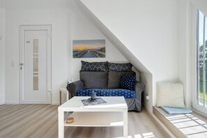 A modern room with a white door, a white table, a grey sofa with patterned cushions, a blue cushion, a white bench, and a painting of a sunset over a bridge. - Apartment 'Villa Wiese  1' with Lake View, Shared Terrace and Wi-Fi (Katzow)