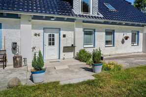 Ein Haus mit weißer Fassade, einer blauen Tür und einem Ziegeldach. Vor dem Haus befinden sich Blumentöpfe.
