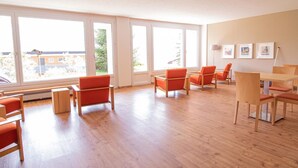Ein geräumiges Zimmer mit großen Fenstern, Holzboden und orangenen Stühlen.