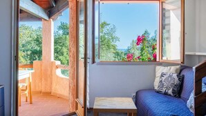 A room with a balcony, a bed, a small table, and a window with a view of greenery and flowers. - Duplex 400 meters from the ocean with a pool in a gated community (Zonza)