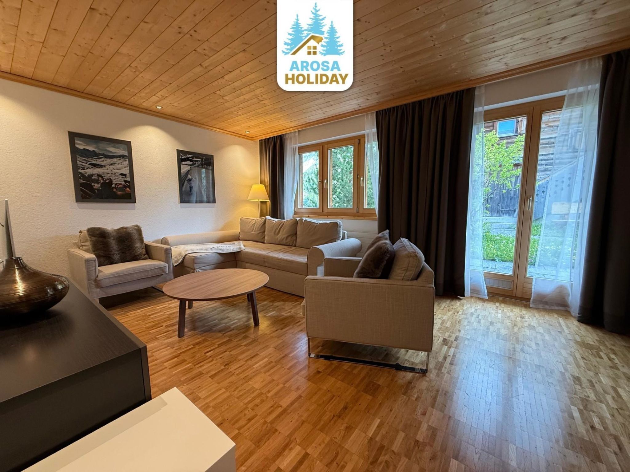 En moderne stue med sofa, lænestole, sofabord og lampe. To vinduer med gardiner, et billede på væggen og et skilt, der viser 'Arosa Holiday'.