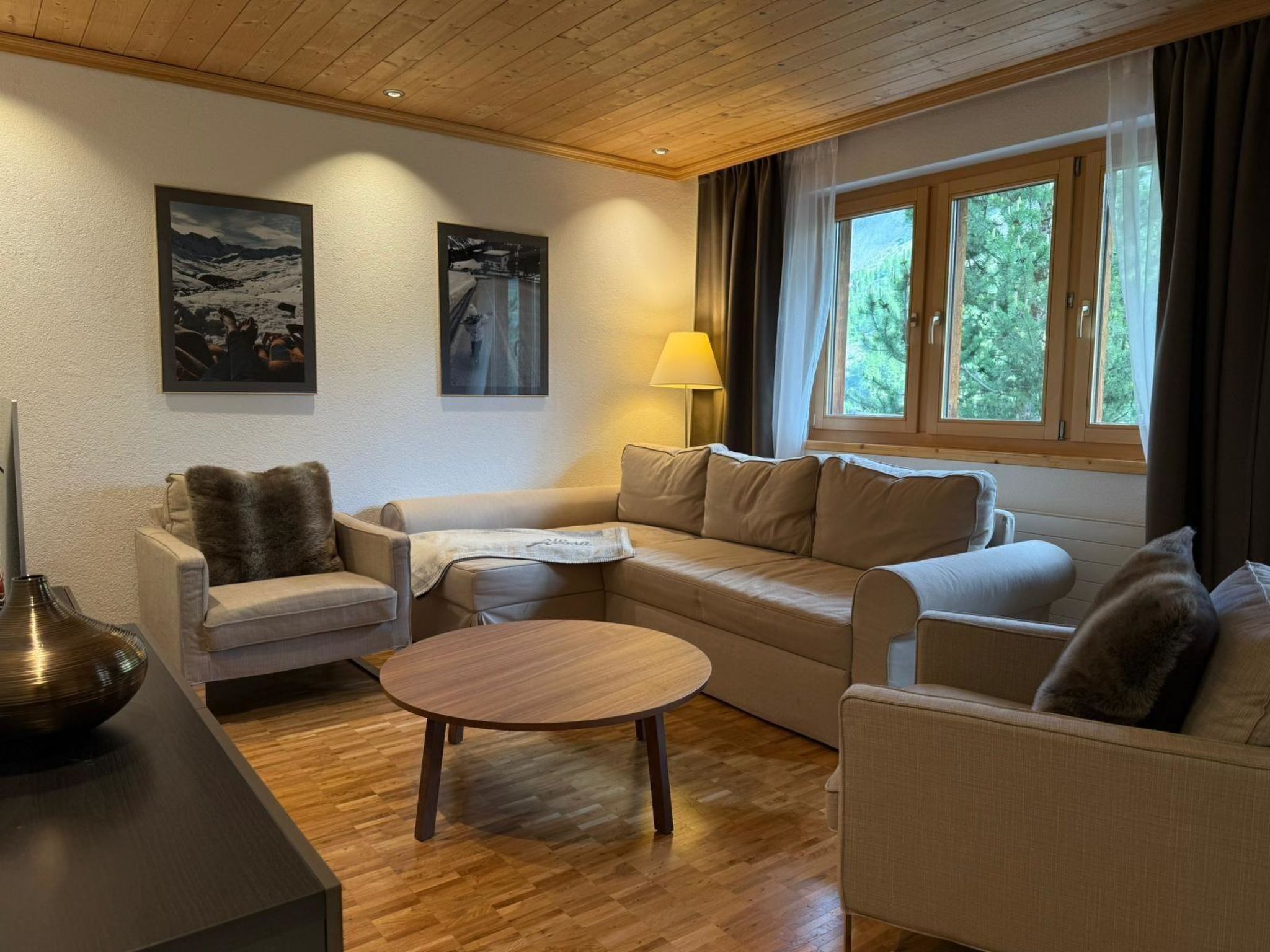 A cozy living room with a sofa, armchairs, a coffee table, and a lamp. There are two windows with curtains, and artwork on the walls.