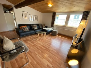 Ein Wohnzimmer mit Holzboden, einem schwarzen Sofa, einem Glastisch, einem gemütlichen Sessel und einem Bild an der Wand.