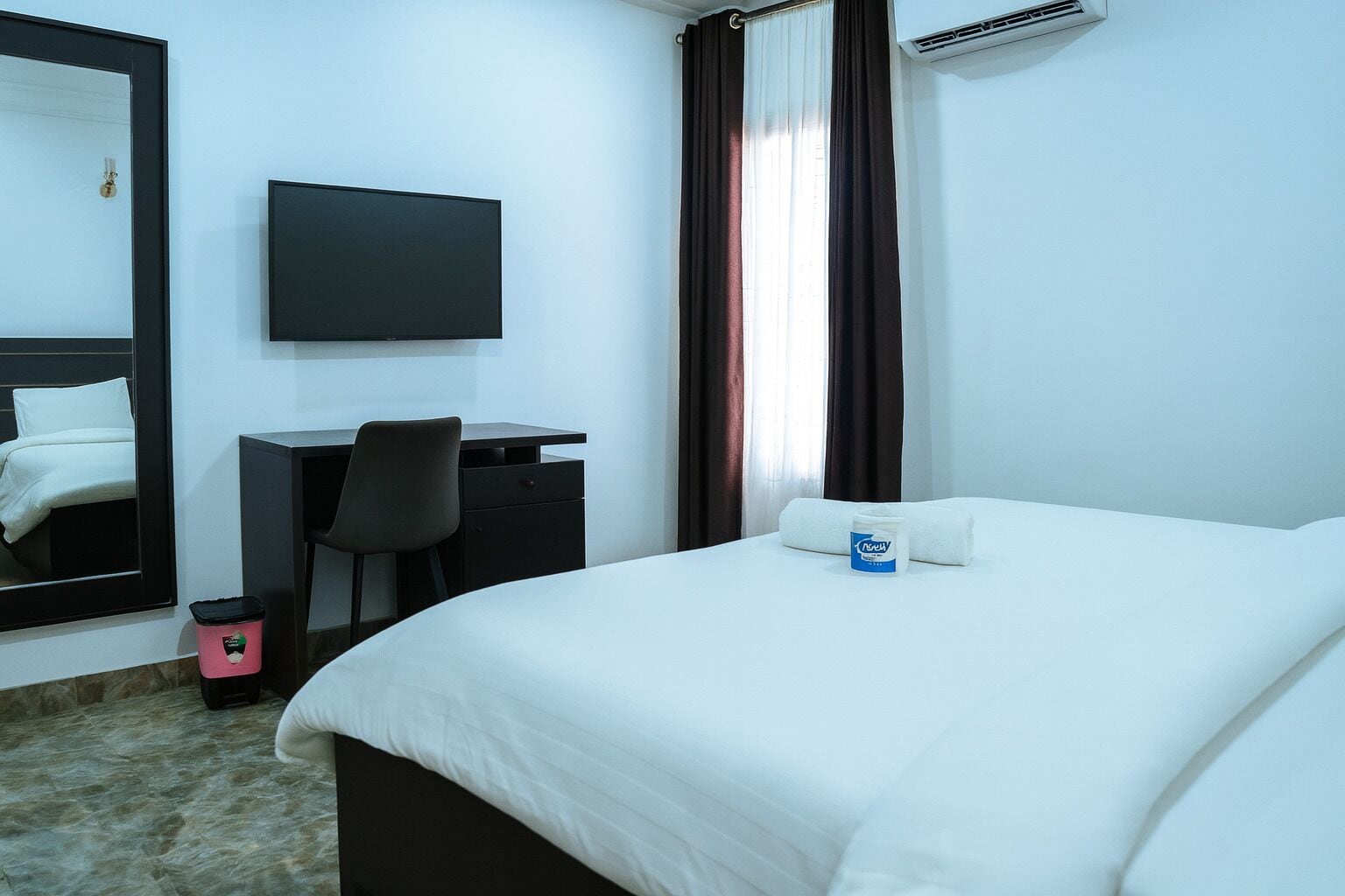 A hotel room with a bed, a desk, a chair, a TV, a window with curtains, and an air conditioning unit.