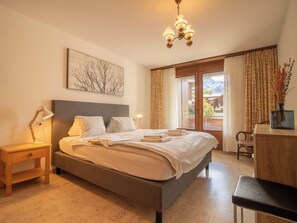 A hotel room with a bed, bedside lamps, a desk, a chair, a window with curtains, and a chandelier. - Brüggli A-24 by Arosa Holiday Appartement/Fewo, Bad, WC, 3-Bettwohnung by Interhome (Arosa)