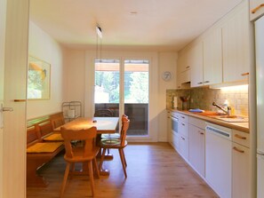 A compact kitchen with wooden cabinets, a dining table with chairs, and a window with a view of greenery. - Furkablick Alice by Arosa Holiday Appartement/Fewo,   Furkablick Alice by Interhome (Arosa)