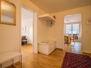 A modern interior with a wooden floor, a white desk, a dining area with chairs, and a large window with a view of trees. - Furkablick Alice by Arosa Holiday Appartement/Fewo,   Furkablick Alice by Interhome (Arosa)