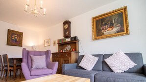 A living room with a wooden coffee table, a purple armchair, a grey sofa, a wooden cabinet, and a wall-mounted clock. - Casa Viktoria A37 by Arosa Holiday Appartement/Fewo, Dusche, WC, 4-Bettwohnung by Interhome (Arosa)