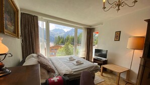A hotel room with a bed, a TV, a desk, a chair, a lamp, and a view of mountains. - Casa Viktoria A37 by Arosa Holiday Appartement/Fewo, Dusche, WC, 4-Bettwohnung by Interhome (Arosa)