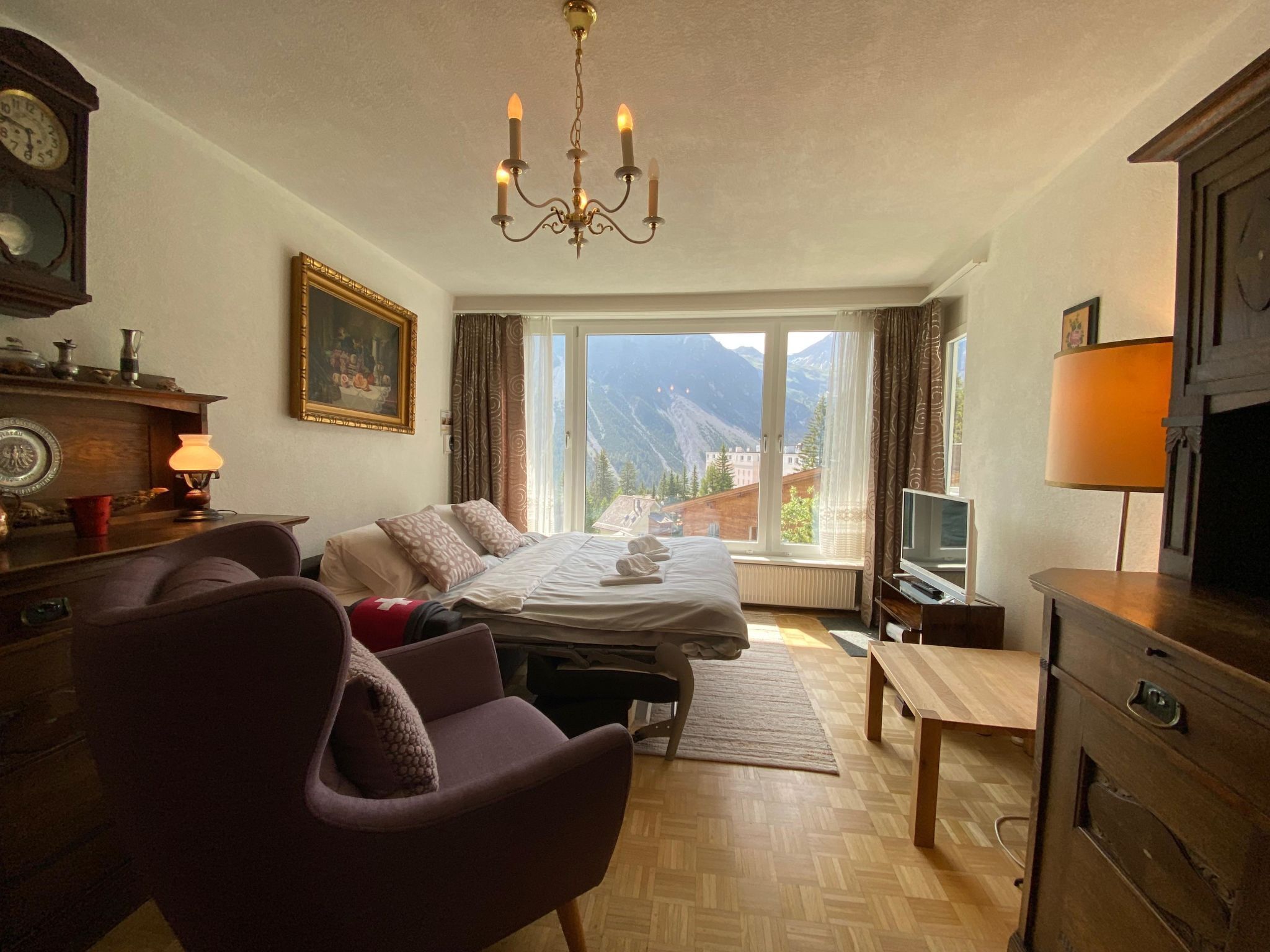 A hotel room with a bed, a desk, a TV, a chair, a lamp, a painting, and a view of mountains.