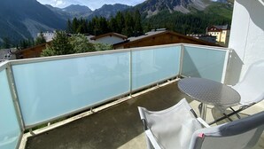 A balcony with a glass railing, a table, and a chair, overlooking a mountainous landscape with houses and trees. - Casa Viktoria A37 by Arosa Holiday Appartement/Fewo, Dusche, WC, 4-Bettwohnung by Interhome (Arosa)