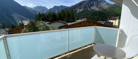 A balcony with a glass railing, a table, and a chair, overlooking a mountainous landscape with houses and trees.