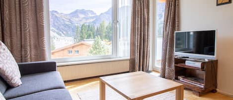 Ein Wohnzimmer mit einer Couch, einem Couchtisch, einem Fernseher auf einem Holzgestell und einem Fenster mit Blick auf Berge.