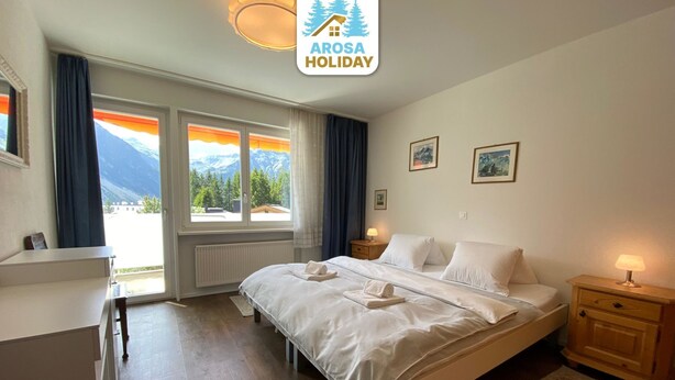A hotel room with a bed, bedside tables, a desk, and a view of mountains. - Casa Viktoria A37 by Arosa Holiday Appartement/Fewo, Dusche, WC, 4-Bettwohnung by Interhome (Arosa)