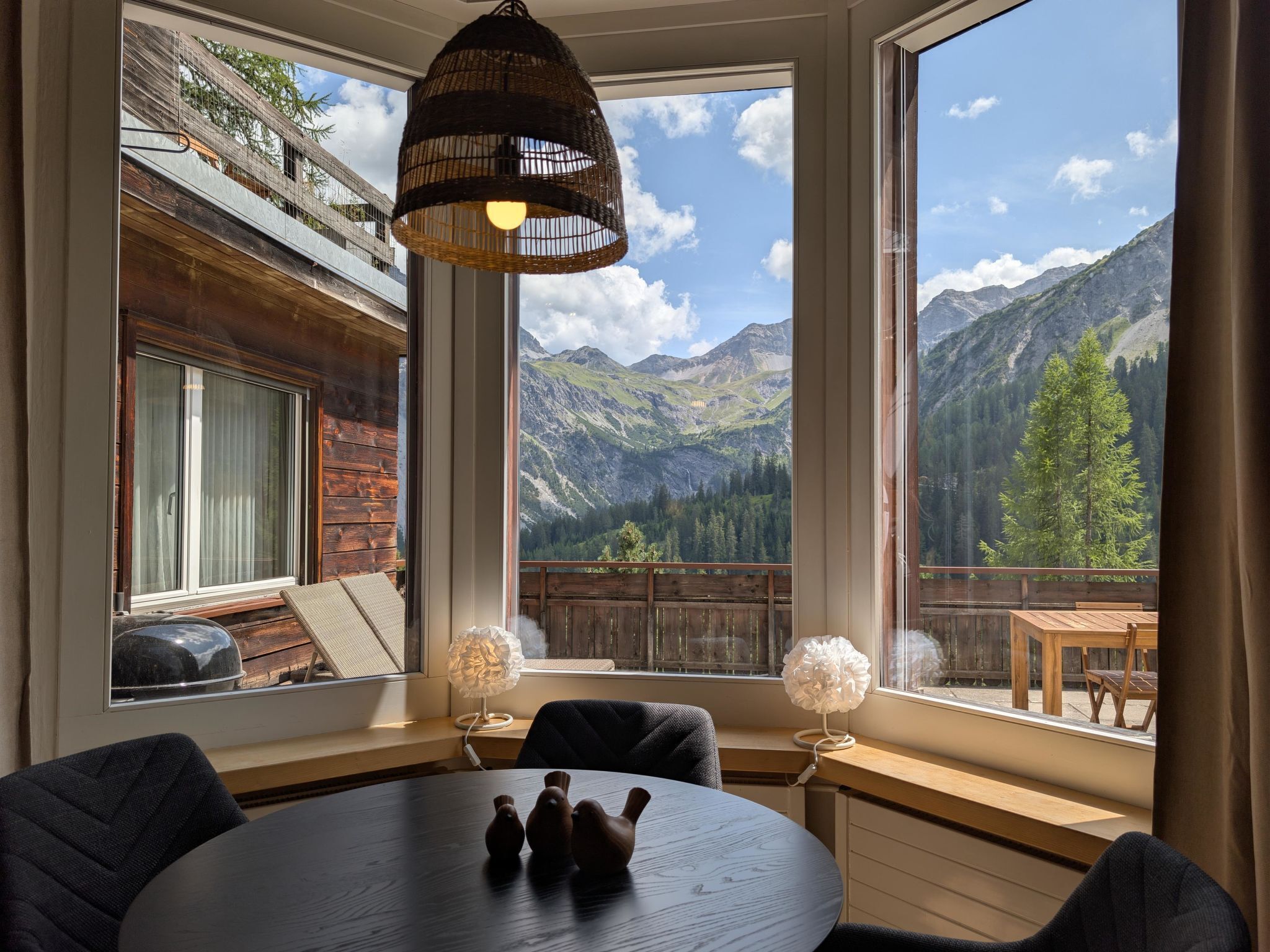 Una habitación con una mesa redonda, sillas, una lámpara colgante y vistas a montañas a través de amplios ventanales.