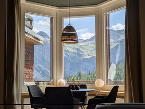 Ein Zimmer mit Tisch und Stühlen, großen Fenstern mit Blick auf die Berge und einer Hängelampe.
