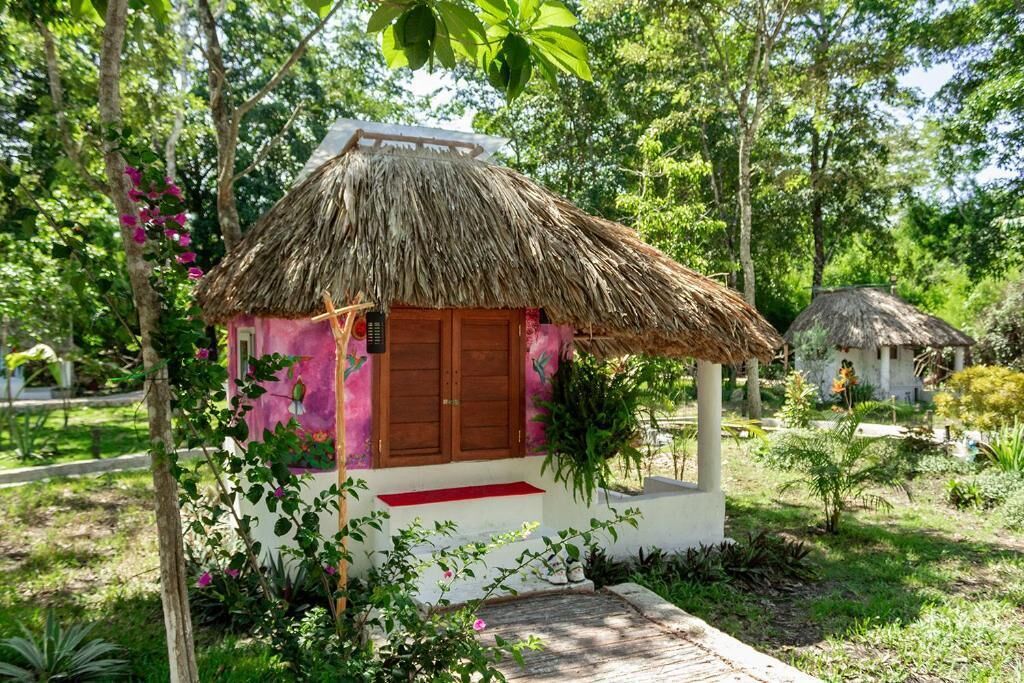 A small, single-story structure with a thatched roof and a red door, surrounded by lush greenery.