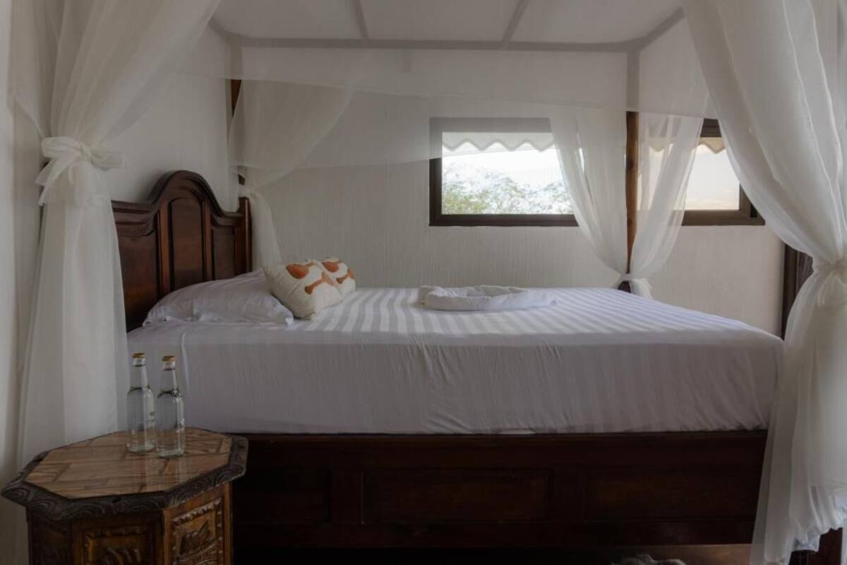 A bedroom with a wooden bed, white bedding, a small bedside table with bottles, and sheer curtains.