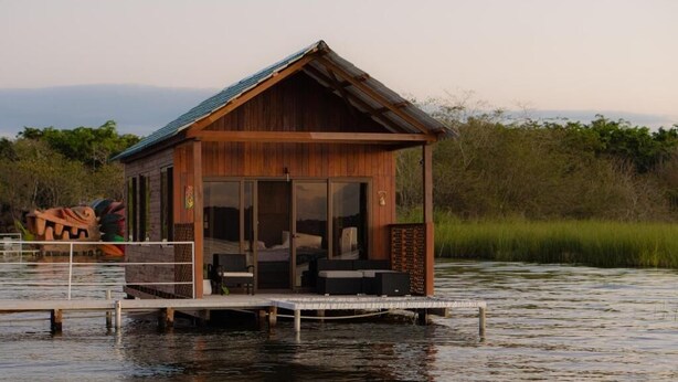 A wooden cabin with a balcony, situated on stilts over water, surrounded by greenery.