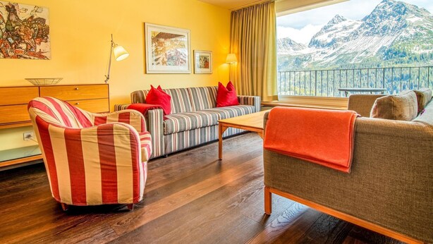 A hotel room with a sofa, armchairs, a wooden coffee table, and a large window with a mountain view. - Casa Linda 8 by Arosa Holiday Appartement/Fewo,  , 7-Bettwohnung Linda 8 by Interhome (Arosa)