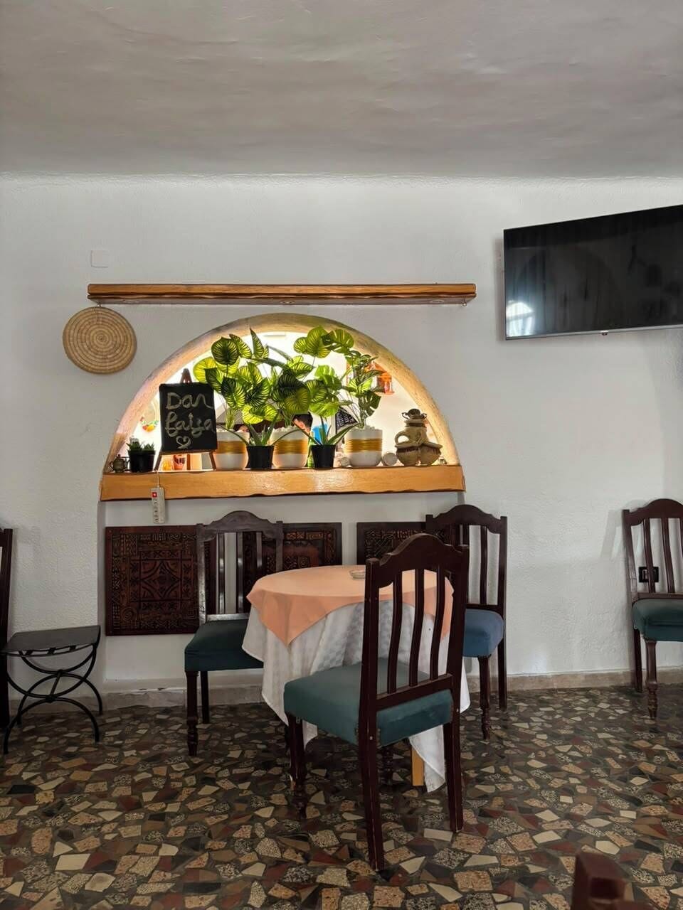 Un espace repas comprenant une table, des chaises, une étagère avec des plantes, un panneau noir et un téléviseur fixé au mur.