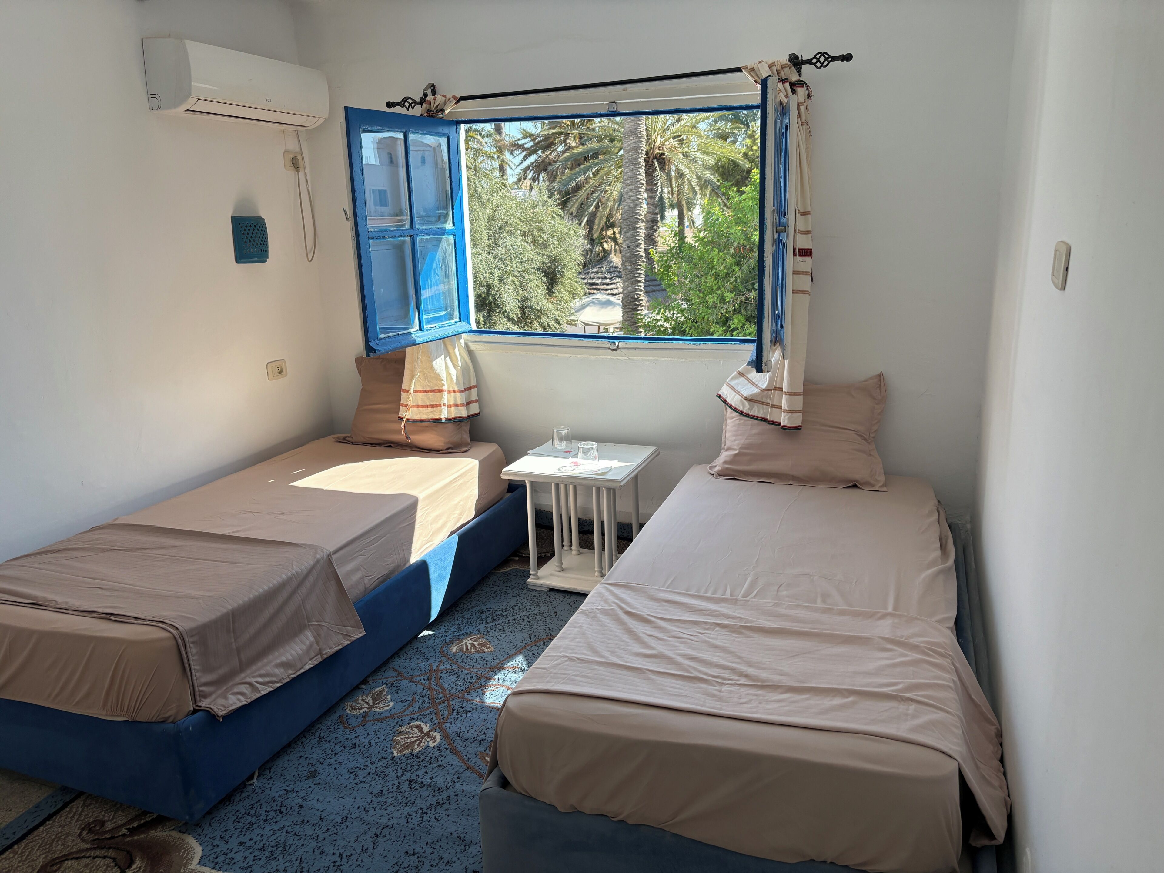 A small room with two single beds, a small table, a window with curtains, and an air conditioning unit.