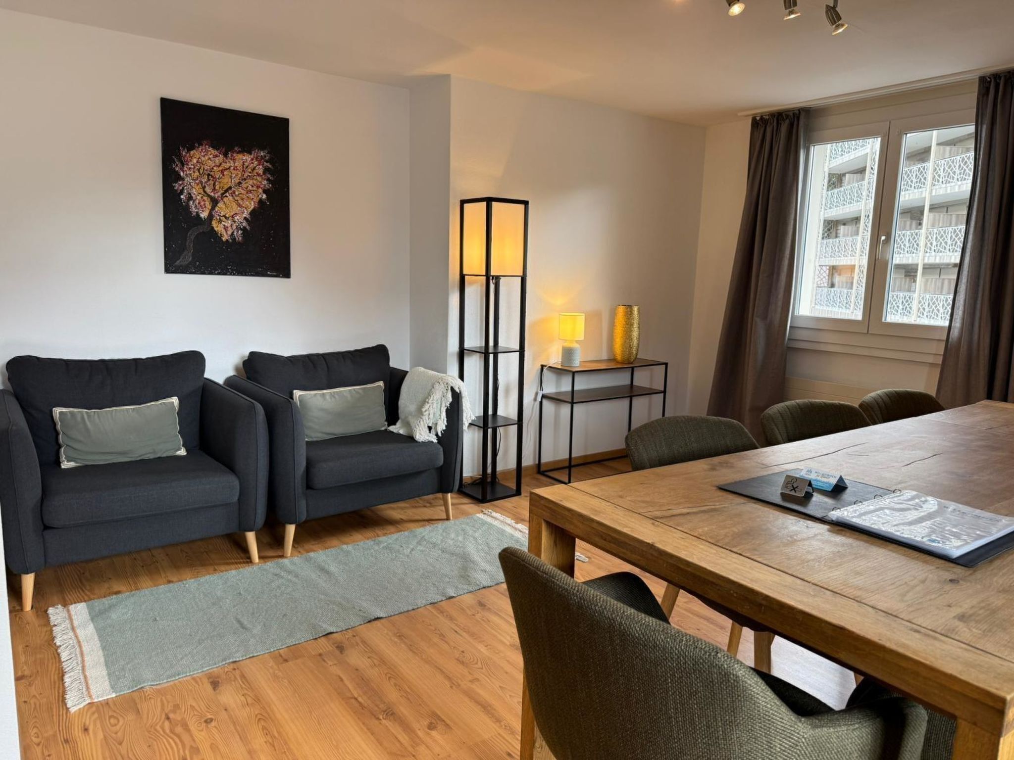 Ein modernes Wohnzimmer mit einer Sofagarnitur, einer Stehlampe, einem Esstisch mit Stühlen und einem großen Fenster mit Vorhängen.