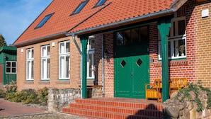 A traditional brick building with a red-tiled roof, green door, and steps leading to a porch with wooden benches.