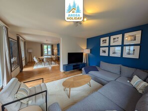 Een moderne woonkamer met een bank, salontafel en televisie. Er is een eethoek met stoelen en een tafel. De kamer heeft een blauwe accentmuur met ingelijste foto's.