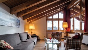 Een gezellige woonkamer met een houten plafond, een bank, een geruite fauteuil, een glazen salontafel en een raam met gordijnen.