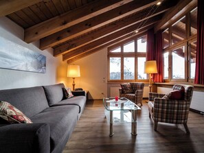 Ein gemütliches Wohnzimmer mit Holzboden, einer Couch, einem karierten Sessel, einem Glastisch und einem Fenster mit Vorhängen.