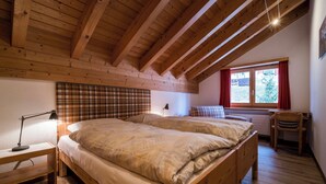 Een slaapkamer met een houten plafond, een bed met een ruitjes hoofdbord, een nachtkastje met een lamp en een raam met gordijnen.