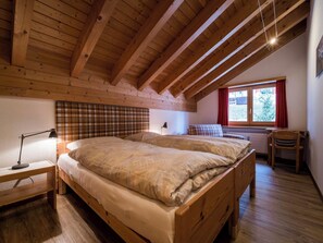 Ein Schlafzimmer mit Holzbalkendecke, einem Bett mit kariertem Kopfteil, einem Nachttisch mit Lampe und einem Fenster mit Vorhängen.